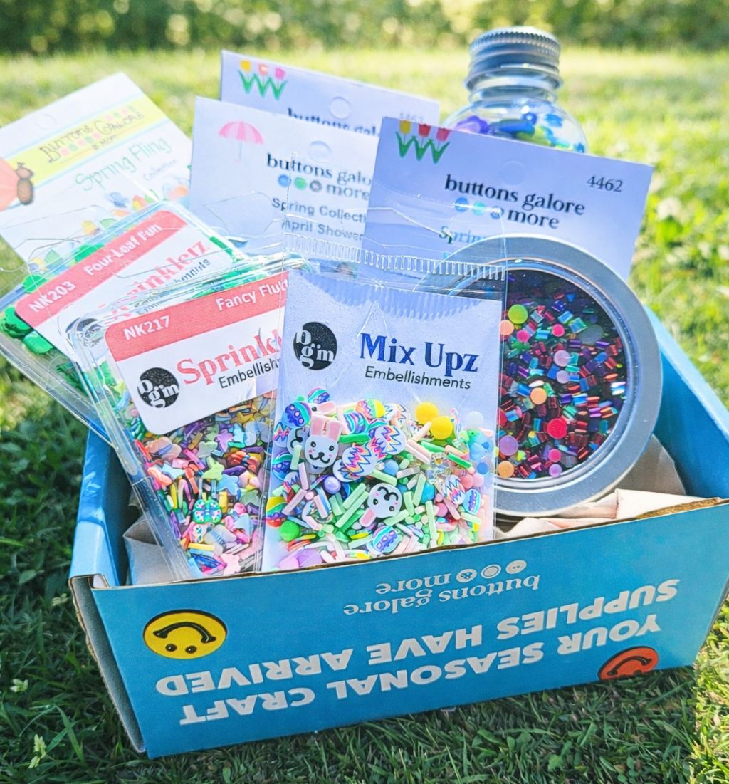 Box of craft supplies including buttons, glitter, and embellishments on grass